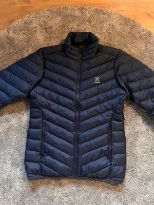 Mörkblå dunjacka från Haglöfs - Mörkblå dunjacka från Haglöfs med hög krage och quiltad design. Jackan har dragkedja framtill, två sidofickor med dragkedja och en liten vit Haglöfs-logga på bröstet. Tillverkad i lätt och värmande material, perfekt för kalla vinterdagar.