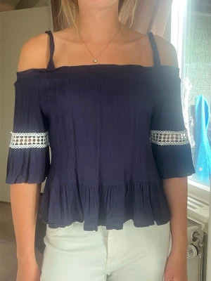 Mörkblå off shoulder topp med spets - Trendig mörkblå off shoulder topp med volangkant och trekvartsärmar. Dekorativ vit spetsdetalj runt ärmarna. Tunn och luftig i materialet, perfekt för sommardagar. Snygg och avslappnad passform med axelband.