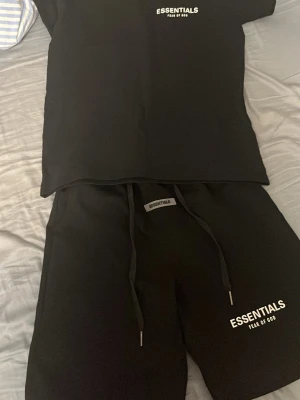 Svart Essentials set med tryck - Snyggt svart set från Essentials Fear of God med t-shirt och shorts. Båda delarna har vitt Essentials-tryck framtill. Shortsen har dragsko i midjan och mjukt material. Perfekt för en avslappnad och trendig look. Pris kan diskuteras vid snabb affär.