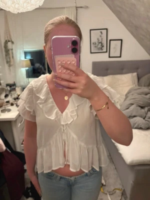 Vit volangblus med kort ärm - Söt vit blus med volangdetaljer längs urringningen och nederkanten. Blusen har korta ärmar och knäppning framtill. Tillverkad i ett tunt, lätt och något transparent material som ger en luftig känsla. Perfekt till jeans eller kjol för en trendig look.