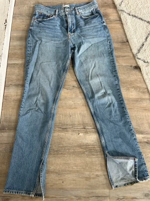 Blå raka jeans med slits  - Jeansen har en ljus tvätt och synliga slitningar vid benslut. Tillverkade i kraftig denim med dragkedja och knappgylf.