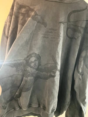 Grå hoodie med ängeltryck från Represent - Säljer en oversized grå hoodie från Represent med unikt tryck av änglar och pilbågar över hela plagget. Hoodien har en tvättad vintage-look, huva och ribbade muddar. Perfekt för dig som gillar streetwear med konstnärlig vibe.