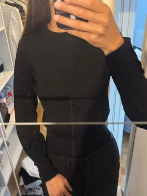 Svart långärmad blus med gulddetaljer - Snygg svart långärmad blus med tight passform och rund halsringning. Ärmarna har dekorativa guldfärgade knappar vid mudden som ger en elegant touch. Perfekt att styla till både jeans och kjol för en trendig look.