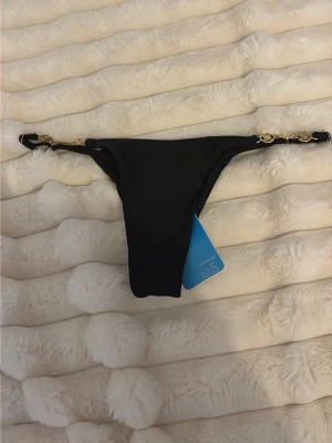 Svarta bikiniunderdel med gulddetaljer - Snygg svart bikiniunderdel från Lindex med guldfärgade kedjedetaljer i sidorna. Klassisk modell med hög skärning, scrunch där bak och liten guldfärgad logga bak. Perfekt för strandhäng eller poolparty.