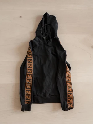 Svart hoodie från Fendi med logoband - Svart hoodie från Fendi med huva och dragsko. Ärmarna har breda bruna band med Fendis ikoniska FF-logga längs hela längden. Tillverkad i mjukt bomullsmaterial och har ribbade muddar vid ärmslut och nederkant. Perfekt för en trendig streetwear-look.