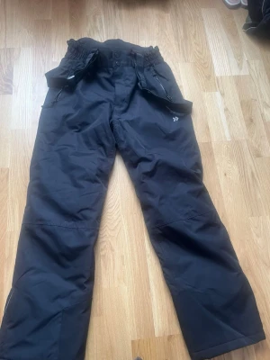 Svarta täckbyxor från 57 Funk - Svarta täckbyxor från 57 Funk med justerbara hängslen och resår i midjan. Byxorna har raka ben och är tillverkade i ett slitstarkt, vattenavvisande material som passar perfekt för vinterns alla äventyr. Snygg och funktionell design för kalla dagar.