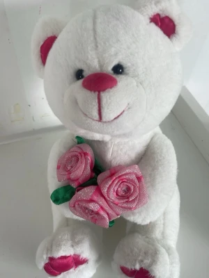 Vit nallebjörn med rosa rosor - Mysig vit nallebjörn i mjuk plysch med rosa detaljer på nos, öron och tassar. Nallen håller tre glittriga rosa tygrosor med gröna blad. Perfekt som present eller dekoration i rummet. En perfekt present till alla hjärtans dag!