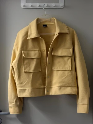 Gul jacka med stora fickor - Stilren gul overshirt jacka i fleece-liknande material med två stora bröstfickor och tryckknappar framtill. Rak passform och klassisk krage ger en modern look. Perfekt att bära som lager över t-shirt eller hoodie.