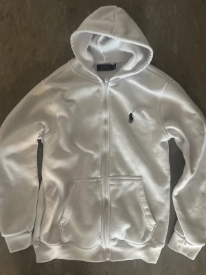Vit hoodie från Polo Ralph Lauren - Snygg vit hoodie med dragkedja från Polo Ralph Lauren. Klassisk design med huva, två fickor framtill och den ikoniska loggan broderad på bröstet. Tillverkad i mjukt material som känns skönt mot huden. Perfekt för en avslappnad och stilren look.