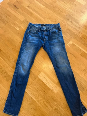 Replay Anbass hyperflex jeans  - Säljer ett par blå jeans från Replay med raka ben och klassisk femficksmodell. Jeansen har snygga slitningar och en tvättad look som ger en avslappnad vibe. Perfekta för dig som gillar en tidlös stil och vill ha ett par jeans som funkar till det mesta