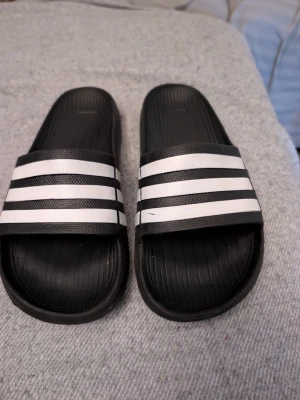 Svarta slides med vita ränder Adidas - Säljer ett par klassiska svarta slides från Adidas med tre vita ränder över foten. Tillverkade i syntetmaterial med räfflad sula för extra grepp. Perfekta för chill dagar eller till stranden. Stl 38