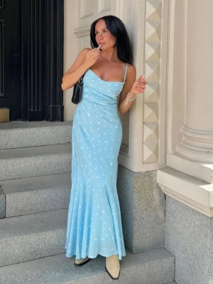 Verona Maxi Dress Adoore - Säljer en ljusblå långklänning med smala axelband och små vita blommor. Klänningen har en figurnära passform upptill och faller mjukt nedtill. Perfekt för sommardagar och har en feminin, elegant look.