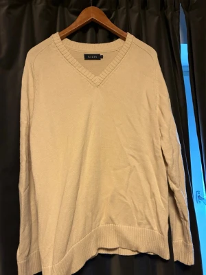 Beige stickad tröja med v-ringning - En stilren beige stickad tröja från Riley med v-ringad hals och ribbade muddar vid ärmslut och nederkant. Perfekt för lager på lager och enkel att matcha med olika outfits. Tillverkad i mjukt material för skön känsla.