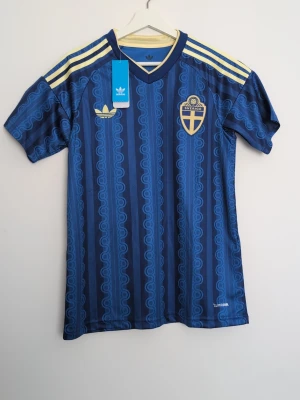 Blå Sverige fotbollströja Adidas - Snygg blå fotbollströja från Adidas med Sveriges landslagsemblem på bröstet och gula detaljer på axlarna. Tröjan har ett unikt mönster i olika blå nyanser, korta ärmar och rund hals. Tillverkad i lätt och funktionellt material, perfekt för träning eller match.