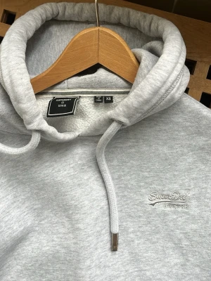 Ljusgrå hoodie från Superdry (XS) - Säljer en ljusgrå hoodie från Superdry i storlek XS. Hoodien har en broderad logga på bröstet och mjukt fleecefoder på insidan. Klassisk passform med ribbade muddar vid ärmslut och nederkant. Perfekt för en chill och avslappnad stil.