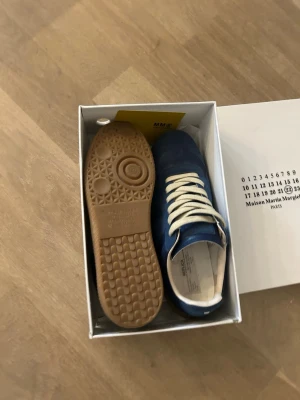 Blå sneakers Maison Margiela - Snygga blå sneakers från Maison Margiela med beige snörning och gummisula. Ovandelen är i en mix av mocka och läder vilket ger en schysst kontrast. Klassisk låg modell som passar till det mesta. Märkeslogga på plösen och insidan.
