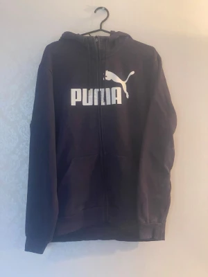 Svart hoodie med dragkedja från Puma - Säljer en svart hoodie från Puma med vit logga och text på bröstet. Hoodien har dragkedja framtill, två fickor och en klassisk huva. Tillverkad i mjukt bomullsmaterial som är skönt att ha på sig. Perfekt för en avslappnad och sportig stil.