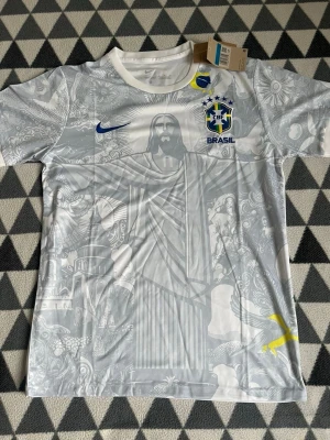 Vit Brasilien fotbollströja Nike M - Säljer en vit Brasilien fotbollströja från Nike i storlek M. Tröjan har ett detaljerat tryck med Kristusstatyn och brasilianska motiv, samt Brasilien-emblem och Nike-logga i blått. Tillverkad i lätt och ventilerande material, perfekt för träning eller match.