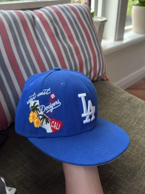 Blå LA Dodgers keps Cali brodyr - Snygg blå keps med LA Dodgers-logga framtill och broderade detaljer med texten 'west coast', blommor, palm och ett rött hjärta med 'CALI' på sidan. Rak skärm och klassisk passform. Perfekt för dig som gillar streetwear och sportig stil.
