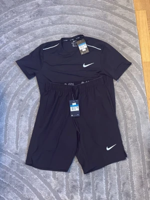 Svart träningsset från Nike - Snyggt svart träningsset från Nike med matchande t-shirt och shorts. Båda plaggen har vita Nike-loggor och är tillverkade i ett lätt, snabbtorkande material som passar perfekt för träning eller löpning. T-shirten har rund hals och shortsen har resår i midjan.