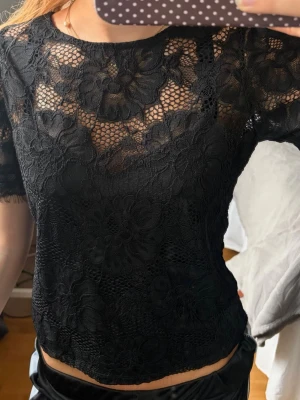 Svart spetsblus med blommönster - Snygg svart blus i genombruten spets med stora blommönster. Kortärmad modell med rund halsringning och transparent material som ger en trendig look. Perfekt att styla med både jeans och kjol för en cool vibe.