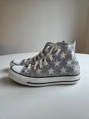 Converse All Star stjärnmönstrade highs - Gråa Converse All Star sneakers med vita stjärnor, vita snören och tåhätta i gummi. Klassisk hög modell med tygmaterial och svart rand på sulan. Perfekta för dig som vill sticka ut med ett coolt mönster.