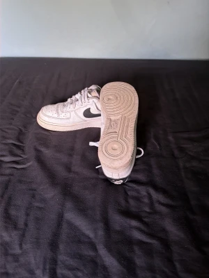 Nike Air Force 1 vita sneakers - Klassiska vita Nike Air Force 1 sneakers med svart swoosh på sidan. Lågt skaft, perforerad tåbox och robust gummisula. Skorna har en tidlös design och passar perfekt till streetwear och casual outfits.