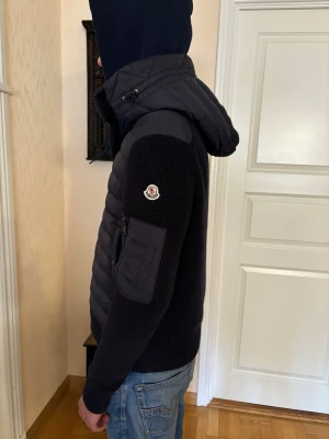 Mörkblå cardigan från Moncler - Snygg mörkblå pufferjacka från Moncler med stickade ärmar och vadderad kropp. Jackan har huva, dragkedja framtill och en ficka med dragkedja på ärmen. Moncler-logga på vänster ärm. Perfekt för kalla vinterdagar.