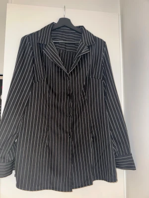 Svart randig skjorta med knappar - Snygg svart skjorta med vita vertikala ränder och klassisk krage. Skjortan har långa ärmar och knäppning framtill. Perfekt för en stilren look.