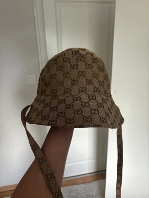 Beige bucket hat från Gucci - Säljer en beige bucket hat från Gucci med klassiskt GG-monogram över hela hatten. Hatten har bred brätte och långa band som kan knytas under hakan. Tillverkad i canvasmaterial och har en liten Gucci-etikett på sidan. Perfekt för dig som vill ha en trendig accessoar.