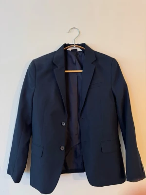 Mörkblå klassisk kavaj HM - Stilren mörkblå kavaj med två knappar framtill och klassisk krage. Kavajen har två fickor med lock och en bröstficka. Passar perfekt till både jeans och kostymbyxor. Materialet är slätt och ger en snygg siluett.
