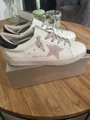 Vita Golden Goose sneakers med stjärna - Snygga vita sneakers från Golden Goose med grå stjärndetalj på sidan och grå skosnören. Skorna har svart hälparti och är tillverkade i läder med slitstark gummisula. Perfekta för dig som gillar en clean och trendig look. Storlek 44 använda nån gång. Pris kan diskuteras bara att höra av sig om man är intresserad. Påse finns till skorna i lådan.