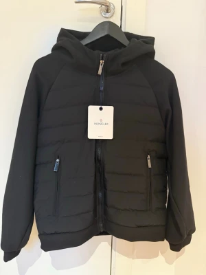Svart pufferjacka från Moncler - Säljer en svart pufferjacka från Moncler med huva och dragkedja framtill. Jackan har Moncler-logga på vänster ärm och två sidofickor med dragkedja. Materialet är en mix av vadderad front och mjuka ärmar, perfekt just nu när våren kommit!