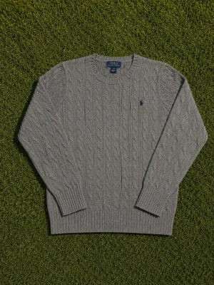 Grå kabelstickad tröja Polo Ralph Lauren - Kabelstickad grå tröja från Polo Ralph Lauren med rund halsringning och diskret broderad logotyp på bröstet. Tröjan har långa ärmar och ribbade muddar vid ärmslut och nederkant. Perfekt för en stilren och klassisk look.