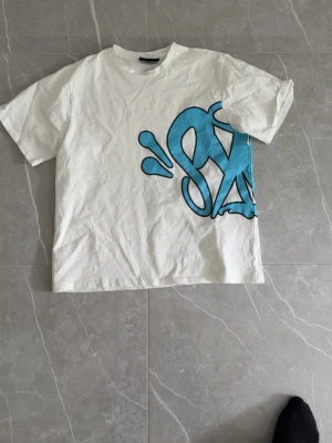 Vit t-shirt med blått graffiti-tryck - Cool vit t-shirt med stort blått graffiti-inspirerat tryck på både fram- och baksidan. T-shirten har klassisk passform och korta ärmar. Perfekt för dig som gillar streetwear och vill sticka ut med en unik design.