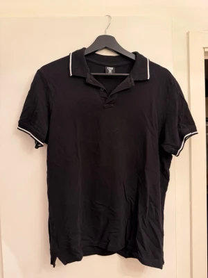 Svart pikétröja från FSBN - Klassisk svart pikétröja från FSBN med vit kant på krage och ärmslut. Slim fit-modell med korta ärmar och knappar vid halsen. Perfekt till jeans eller shorts för en clean look.