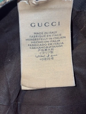 Gucci beige keps med GG-mönster - Säljer en beige keps från Gucci med det klassiska GG-monogrammet i brunt över hela kepsen. Kepsen har böjd skärm och detaljer i grönt och rött på sidorna. Tillverkad i Italien och i slitstarkt tygmaterial. Perfekt för dig som vill ha en ikonisk och trendig accessoar.