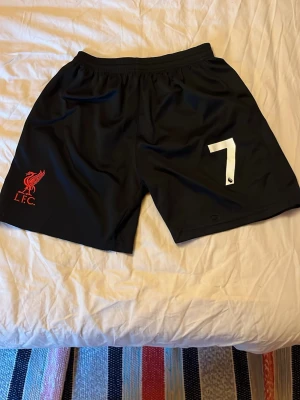 Liverpool FC svarta shorts nr 7 - Svarta sportshorts från Liverpool FC med vitt nummer 7 på höger ben och röd L.F.C.-logga på vänster ben. Elastisk midja för skön passform. Perfekta för fotboll eller träning, tillverkade i lätt och snabbtorkande polyester.