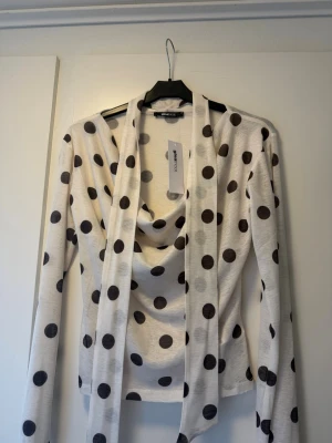 Prickig långärmad topp Gina Tricot - Säljer en vit långärmad topp från Gina Tricot med stora svarta prickar. Toppen har en snygg draperad ringning och långa band framtill som kan knytas på olika sätt. Perfekt för dig som gillar trendiga detaljer och mönster.