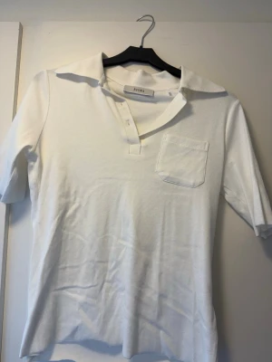 Vit henley-skjorta från Avora - Klassisk vit henley-skjorta från Avora med kort ärm, krage och bröstficka. Skjortan har knappar framtill och är tillverkad i ett mjukt bomullstyg. Perfekt för en clean och stilren look.