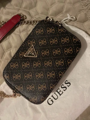 Svart och brun handväska från Guess - Snygg handväska från Guess i svart konstläder med brunt monogrammönster och guldfärgade detaljer. Väskan har en kedjerem med röd axelrem och dubbla dragkedjor upptill. Perfekt storlek för det viktigaste och en trendig accessoar till din outfit.