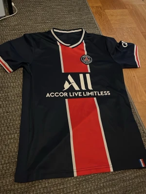 PSG matchtröja mörkblå/röd - Säljer en snygg PSG matchtröja i mörkblått med bred röd rand och vita detaljer. Tröjan har klubbmärke på bröstet och sponsorlogga framtill. Korta ärmar med röd, vit och blå kant. Perfekt för fotbollsfans!