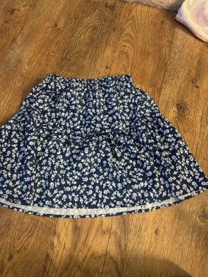Blå blommig kortkjol med volang - Söt blå kortkjol med vitt blommönster och volangdetaljer. Kjolen har en avslappnad passform och resår i midjan för extra komfort. Perfekt för dig som gillar en feminin och lekfull stil.