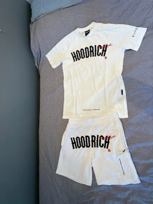 Vit HOODRICH t-shirt med tryck - Säljer ett vitt set från Hoodrich med matchande t-shirt och shorts. Båda plaggen har stort svart HOODRICH-tryck med röd detalj framtill. T-shirten har rund hals och korta ärmar, shortsen har resår i midjan och fickor. Perfekt för en avslappnad streetwear-look.