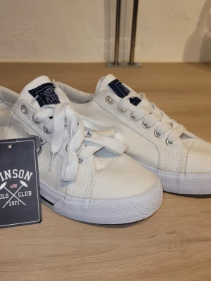 Vita låga sneakers från Vinson Polo Club - Snygga vita sneakers i canvas från Vinson Polo Club med klassisk låg profil. Skorna har vita snören, silverfärgade öljetter och logga på sidan samt plös. Perfekta för en clean och sportig stil.