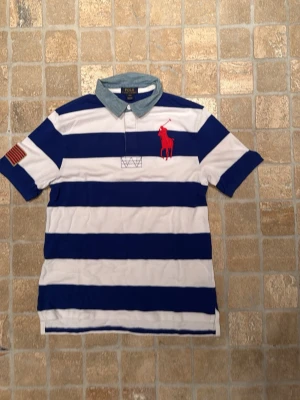 Randig pikétröja från Polo Ralph Lauren - Säljer en snygg pikétröja från Polo Ralph Lauren med breda blå och vita ränder, röd logga på bröstet och denimliknande krage. Tröjan har korta ärmar och klassisk knappslå. Perfekt för en avslappnad men stilren look.