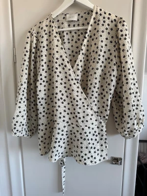 Blommig omlottblus från Neo Noir - Söt omlottblus från Neo Noir i vit med små svarta blommor. Blusen har trekvartsärm med lätt puff och knytband i midjan. Materialet är strukturerat och ger en trendig look. Perfekt att styla med jeans eller kjol.