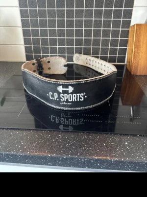 Svart träningsbälte från C.P. Sports - Snyggt svart träningsbälte från C.P. Sports med vit logga och kontrastsömmar. Bältet är tillverkat i skinn och har en bred, vadderad design som ger extra stöd vid tunga lyft. Perfekt för gymmet och styrketräning.