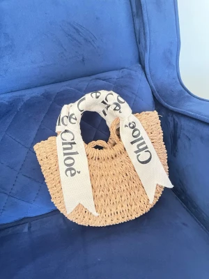 Stråväska  med band - Snygg handväska från Chloé i naturfärgat flätat stråmaterial. Väskan har rundad form och handtag dekorerade med breda vita band med Chloé-logga i svart. Perfekt accessoar för en trendig look.