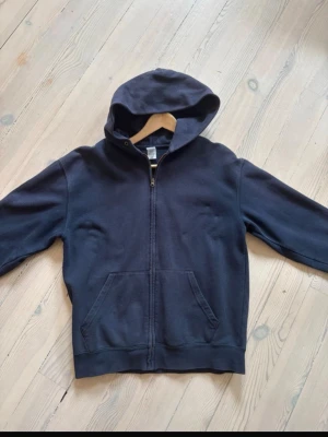 Mörkblå zip-hoodie från fruit of the loom - Mörkblå hoodie med dragkedja från Fruit of the Loom. Klassisk modell med huva och två stora framfickor. Tillverkad i mjukt bomullsmaterial, perfekt för en avslappnad stil. Enkel och stilren design utan tryck.